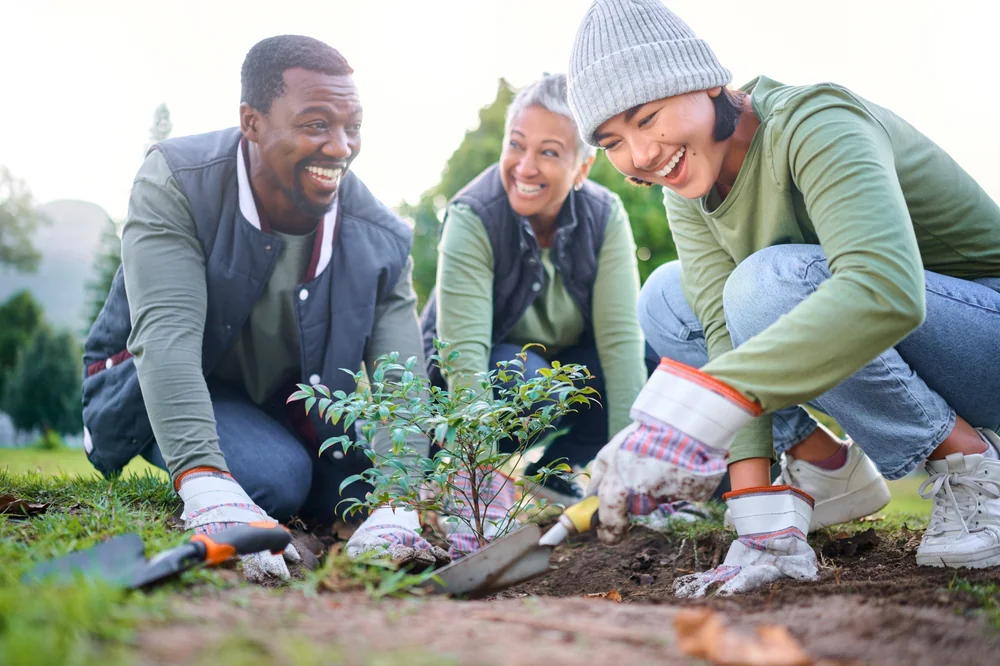 Three people kneeling on grass, smiling and planting a small tree together. They wear gloves and casual outdoor clothing, working as a team in a green park or garden setting.